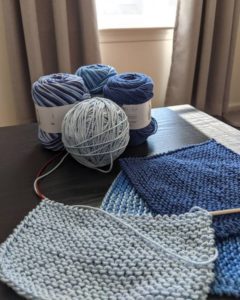 Four skeins of blue yarn on a table with 3 knit washcloths, one of which is in progress. A sunny window with grey curtains shines in the background.