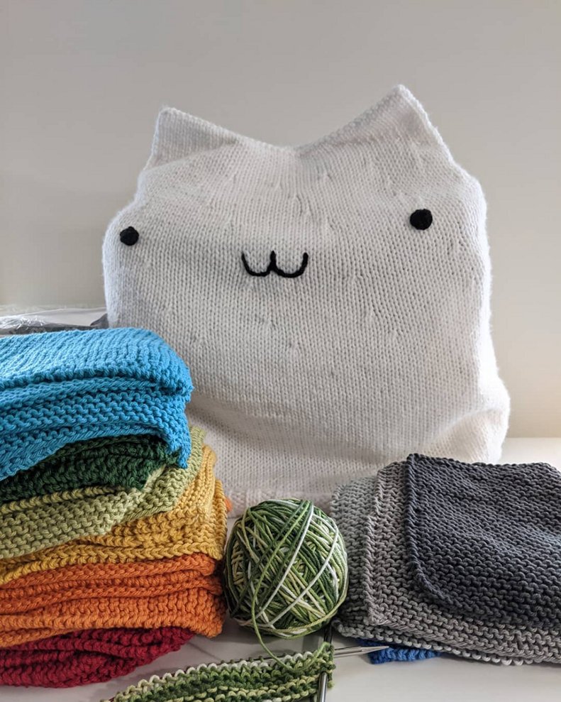 Two piles of colorful knitted washcloths stacked in front of a white knitted Bongo Cat cover which protects the sewing machine beneath it.