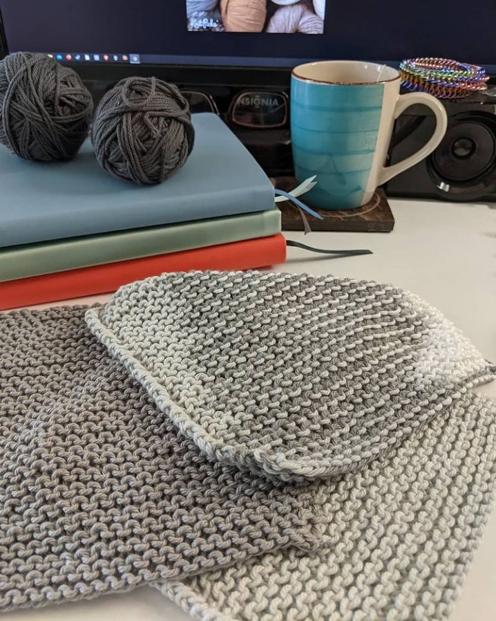 Photo: A work desk, with colorful journals stacked in front of a flatscreen monitor and next to a blue coffee mug, and a stack of washcloths in various shades of gray in the for