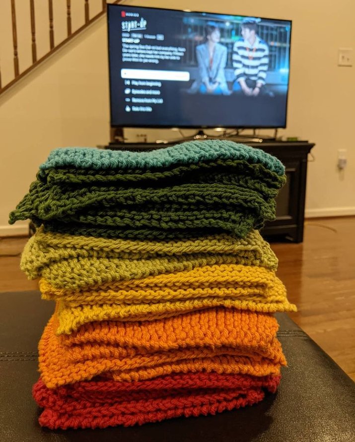 A stack of knit cloths, blue to red, stacked on a surface in front of a flat screen TV showing the Netflix interface, with the show "Start Up" on the screen.