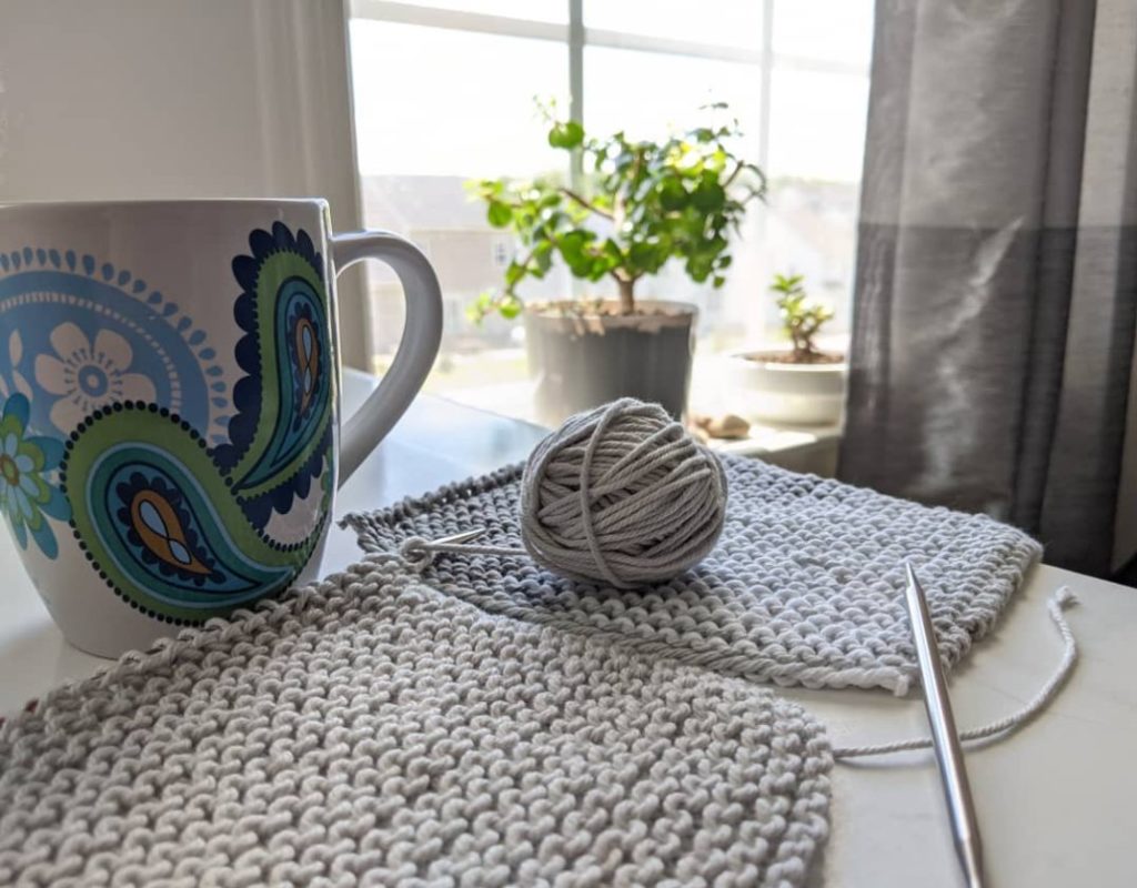 Grey cotton knitted washcloths in progress on a table, next to a painted coffee cup, in front of a sunny window with plants.