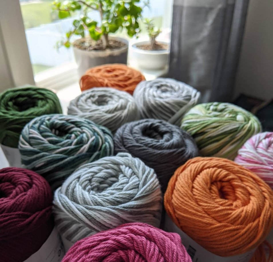 Colorful cotton yarn skeins arranged on a table in front of a sunny window with plants.