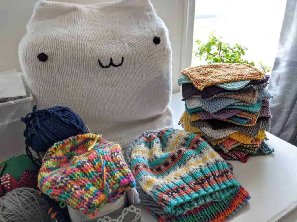 Lots of colorful knitted items, some still in progress, sitting in piles on a table, in front of a sewing machine covered by a knitted Bongo Cat cover, with a sunny plant filled window in the background.