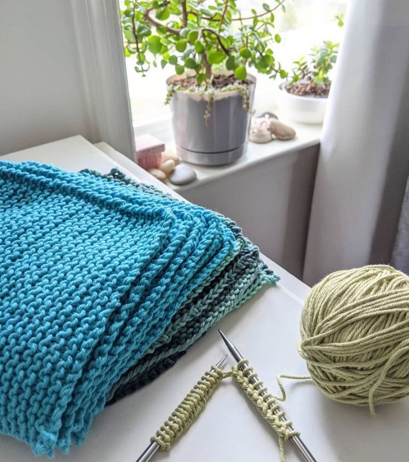 A stack of hand knit washcloths in blues and greens, with one still on knitting needles, sitting near a sunny window with leafy green plants on the sill.