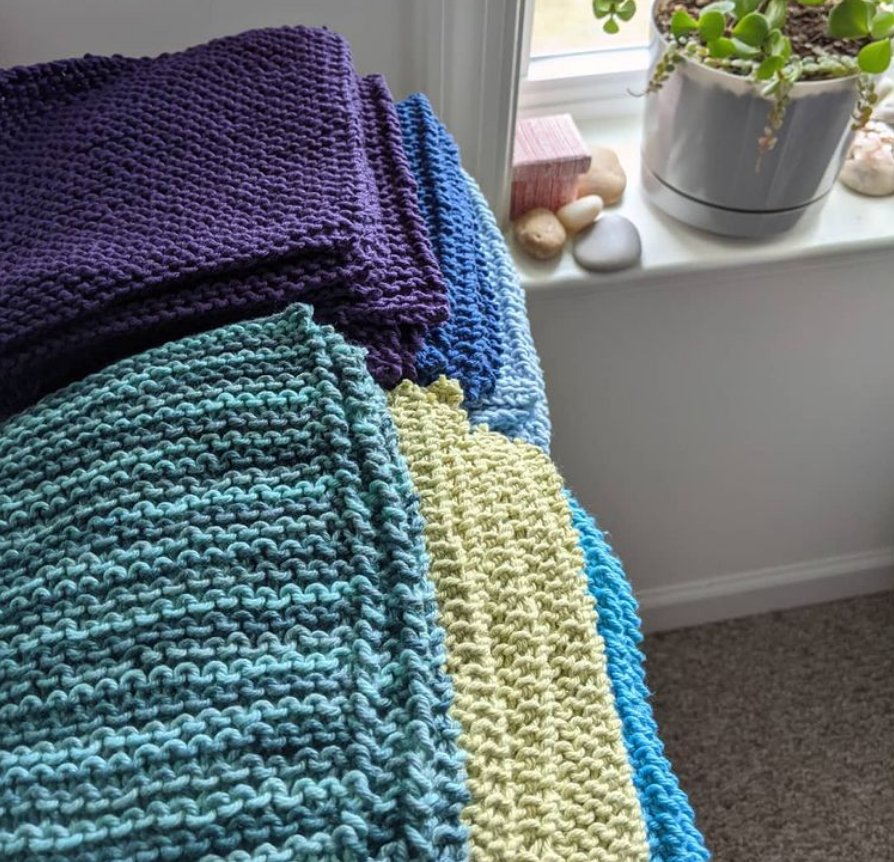 A stack of hand knit washcloths in blues, greens, and purples, sitting near a sunny window with leafy green plants on the sill.