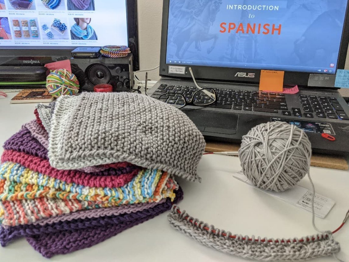 A desktop with laptop and monitor, and a colorful stack of dishcloths, with one cloth on circular needles.