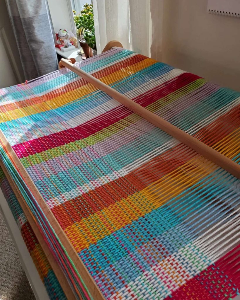 Loom WIP - 32" wide warp of blue, pink, yellow, lavender, white and a hint of green. The variegated weft yarn includes most of the same colors, and part of this photo is illuminated by limited sunlight filtering through the sheer curtains.