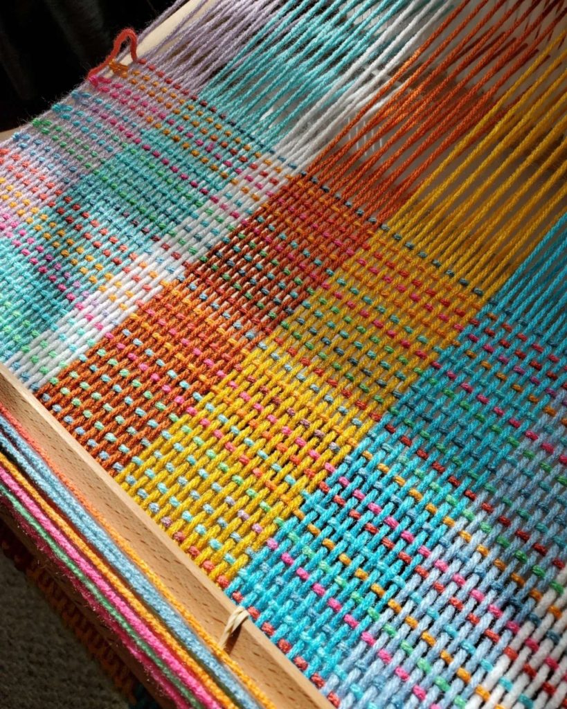 Loom WIP - 32" wide warp of blue, pink, yellow, lavender, white and a hint of green. The variegated weft yarn includes most of the same colors, and part of this photo is illuminated by limited sunlight filtering through the sheer curtains.